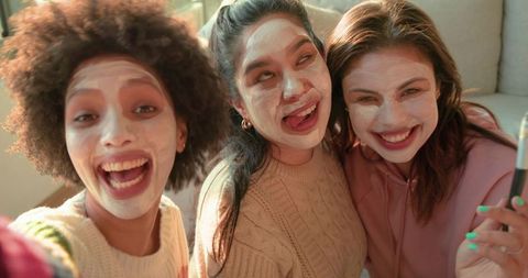 Diverse women taking selfie with face masks on sofa laughing, enjoying skincare selfcare