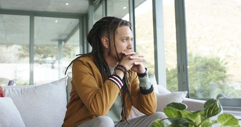 Thoughtful african american man sitting on sofa gazing out window in modern living room