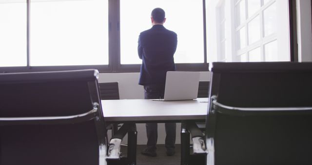 A businessman in a suit is standing and looking out of an office window. An empty conference room table and chairs are visible, with a laptop on the table. Suitable for themes related to business, professional life, decision-making, and corporate environments.