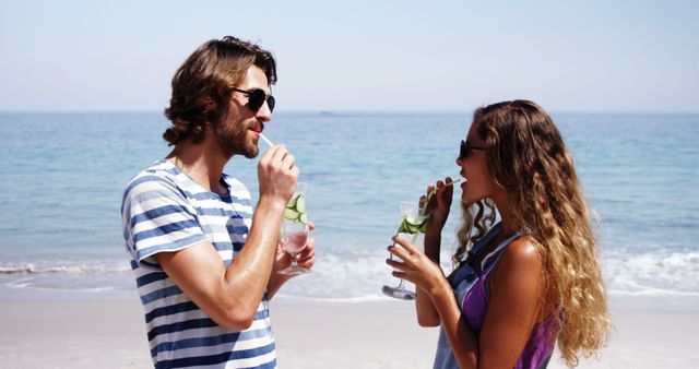 Young couple standing at beach, drinking beverages with garnishes, appearing relaxed. Ideal for promoting summer vacations, beach holidays, travel experiences, and lifestyle blogs.