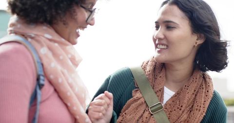 Two women smiling and chatting on urban street wearing knit scarves and casual bags