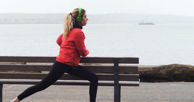 A young Caucasian woman jogs by the seaside, wearing headphones and athletic gear, with copy space. Her focused expression and active posture convey a commitment to fitness and a healthy lifestyle.