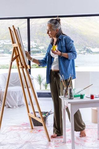 Middle-aged painter working at easel in coastal studio barefoot holding yellow paint jar