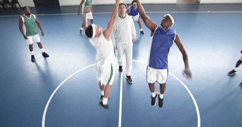 Diverse male athletes jumping for basketball tipoff at gym