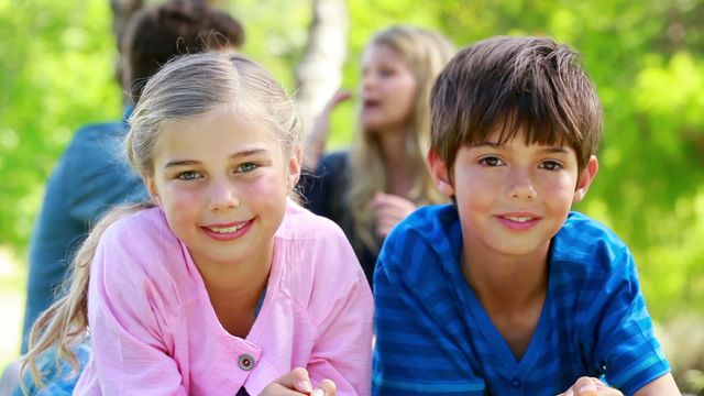 Two children happily posing outdoors with their parents chatting in the background. Suitable for depicting family fun, outdoor activities, and carefree childhood moments. Ideal for parenting articles, family-oriented websites, and advertising related to children's products.