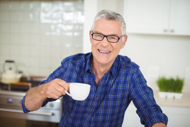 Senior man in plaid shirt and glasses enjoying a cup of tea in a modern kitchen. Ideal for use in articles or advertisements related to senior living, healthy lifestyle, home comfort, and morning routines.