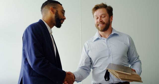 Two professional men are shaking hands in an office environment. One man is holding folders, suggesting they are engaging in a successful business agreement or partnership. This can be used in articles or marketing materials related to business success, professional engagements, teamwork, partnerships, and collaboration in corporate environments.