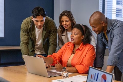 Diverse team collaborating around laptop with presenter pointing at data and charts
