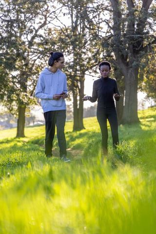 Diverse couple walking sunlit park trail, chatting and using smartphone tracking fitness