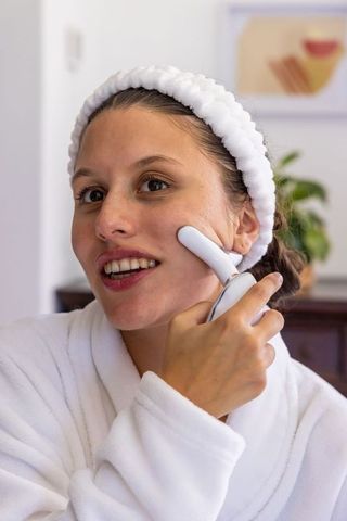 African american woman applying facial device while wearing bathrobe and headband