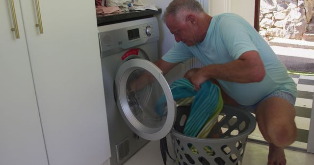 Senior man crouching down while loading clothes into a front-loading washing machine. This image is useful for content related to home life, daily routines, household chores, or advertising home appliances and laundry services.