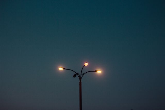 Street light with bright bulbs glowing against a clear evening sky. Suitable for themes of urban tranquility, minimalism, and public infrastructure. Ideal for illustrating concepts related to city life, twilight, and outdoor lighting.