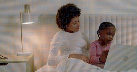 Mother and daughter using laptop on bed in cozy bedroom with warm lamp light