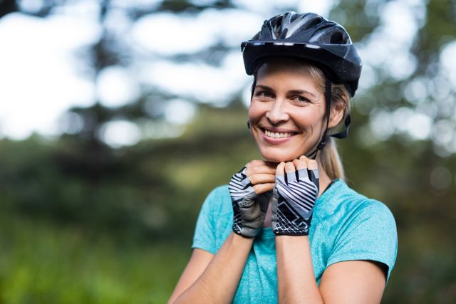 Woman smiling while wearing a bicycle helmet and cycling gloves, standing outdoors with a nature background. Ideal for promoting outdoor activities, fitness, healthy lifestyle, and safety gear. Suitable for use in advertisements, blogs, and articles related to cycling, exercise, and women's sports.
