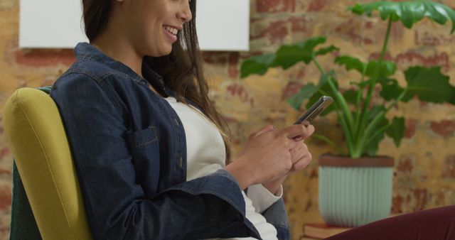 Smiling woman in a denim jacket while using a smartphone in a cozy modern apartment. Could be used for themes like mobile technology, communication, social media interaction, or relaxed home lifestyle.