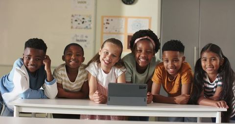Diverse Elementary Students Leaning Over Tablet, Collaborating and Smiling in Classroom
