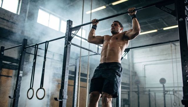 Performing shirtless pull-ups in industrial gym with rings, rope and chalk dust training