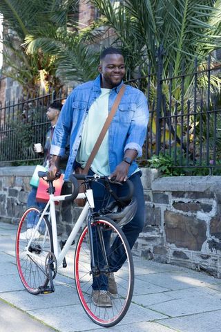 Smiling african american commuter pushing bike along urban sidewalk with helmet and denim