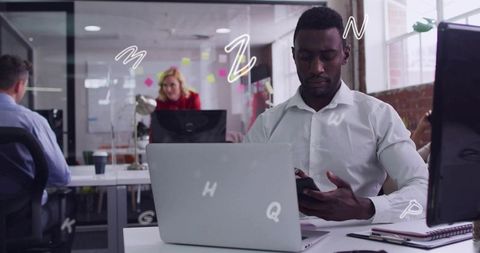 Man using smartphone while working on laptop in modern open-plan office with coworkers
