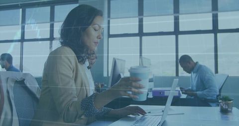 Focused businesswoman typing on laptop while holding takeaway coffee in open-plan office
