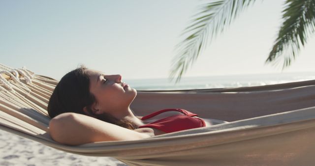 Young woman lounging on a hammock at a sunlit tropical beach, soaking up the tranquility. Ideal for use in travel promotion, vacation packages, lifestyle blogs, and advertisements conveying relaxation and leisure.
