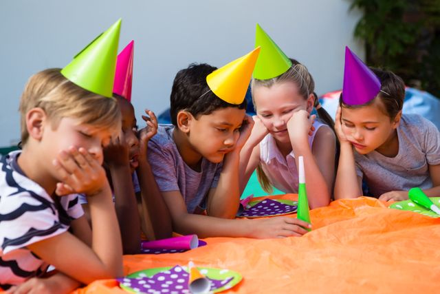 Children sitting at a table during a birthday party, wearing colorful party hats and looking bored. This image can be used to depict childhood emotions, party planning, or the importance of engaging activities at events.