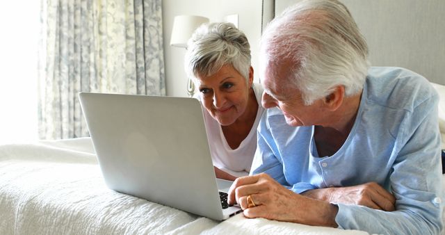 Elderly couple is relaxing in their home bedroom and using a laptop together. This image is perfect for illustrating themes of senior citizens, active aging, technology, connecting with family, and computer literacy among older adults. Suitable for use in articles, blogs, websites, and promotional materials centering on elderly lifestyle, retirement, tech-savvy seniors, and online connectivity.