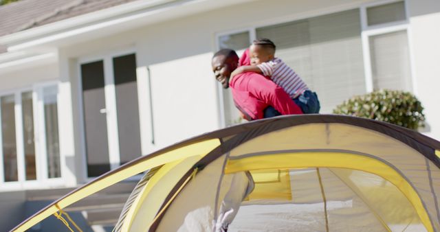 Father enjoying a playful moment with his child by giving a piggyback ride next to a tent in the backyard. Great for depicting family bonding, outdoor activities, parenting moments, and joyful memories.