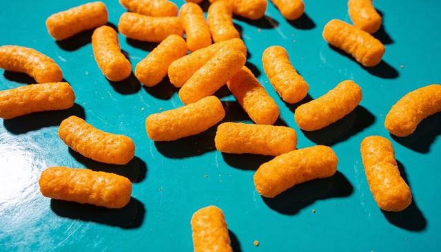 Scattering crunchy cheese puffs on teal surface with crumbs, glossy reflections, shadows
