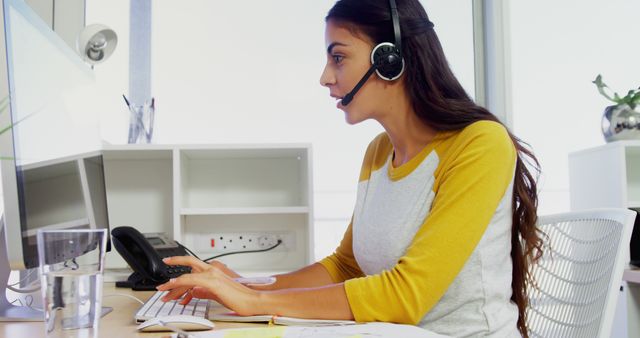 A young Caucasian woman works diligently at her computer while wearing a headset, assisting customers in a call center environment. Her focused demeanor underscores the importance of customer support roles in maintaining client satisfaction.