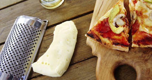 Pizza slice with mushrooms and melted cheese alongside a block of cheese and a metal grater on a wooden table creates a homely and inviting scene, perfect for illustrating food blogs, Italian cuisine recipes, kitchen-related advertisements, or cooking tutorials.