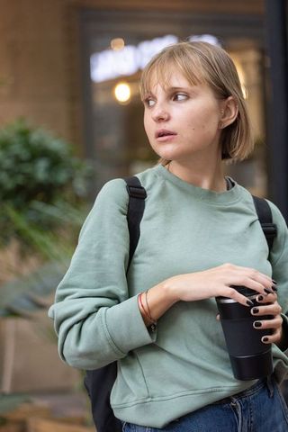Young woman holding matte black travel mug wearing sage sweatshirt in urban cafe scene