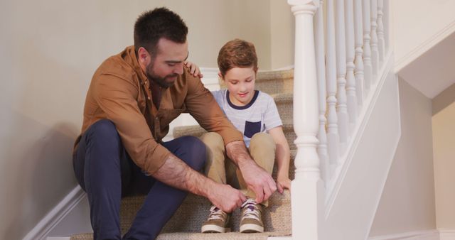 Father showing son how to tie shoes while sitting on stairs in bright home environment. Perfect for family bonding, parenting, and educational themes. Ideal for use in articles, advertisements, or websites centered on child development, family roles, or home life.