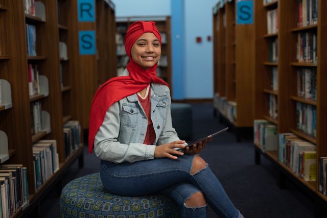 Asian female student wearing a red hijab and denim jacket is studying in a library, sitting on a seat between bookshelves and using a tablet computer. This image is ideal for educational content, technology in education, diversity in learning environments, and promoting digital literacy.