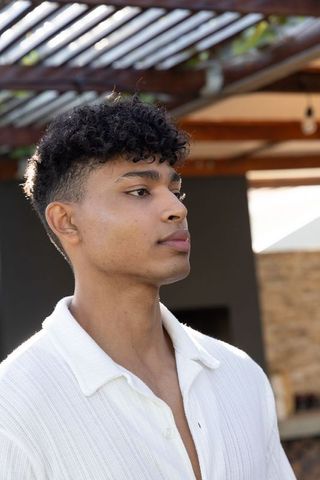 Sunlit african american man standing under pergola on patio wearing white collared shirt