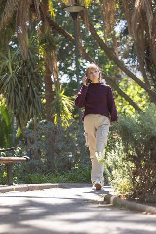 Young woman walking through sunlit park talking on smartphone near bench and palms