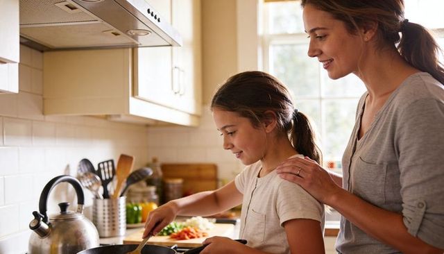 Mother guiding daughter stirring pan at stove in sunlit kitchen, family cooking bond