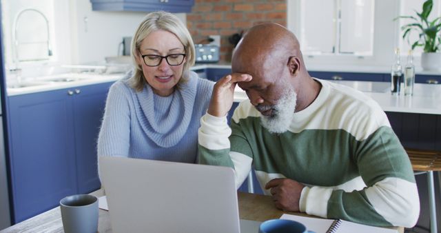 Stressed senior diverse couple in kitchen sitting at table, using laptop. retirement lifestyle, at home with technology.