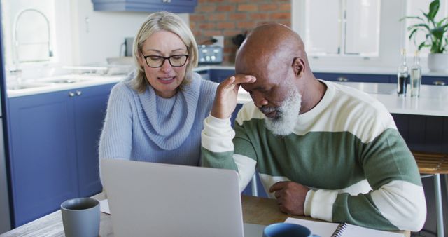 Stressed senior diverse couple in kitchen sitting at table, using laptop - Download Free Stock Photos Pikwizard.com