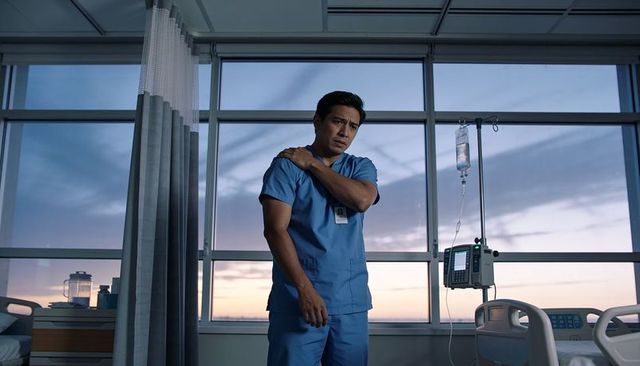 Night shift nurse standing by hospital window touching shoulder, iv pole and empty bed
