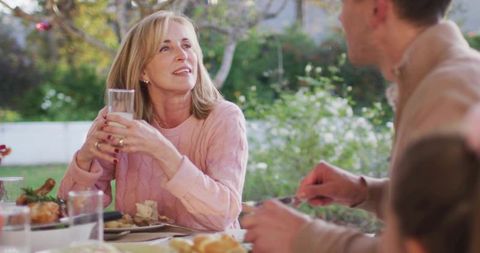 Mature woman holding glass smiling and gazing during outdoor family meal on sunlit patio