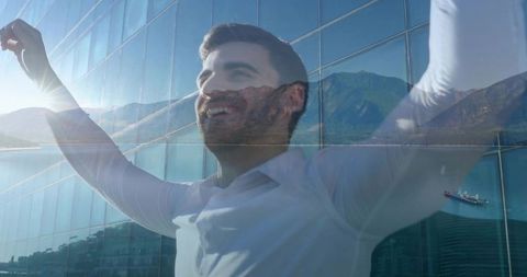 Triumphant businessman raising arms with coastal mountain reflection on glass facade