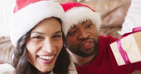Diverse couple wearing santa hats exchanging gift with pink ribbon on couch, cozy holiday moment