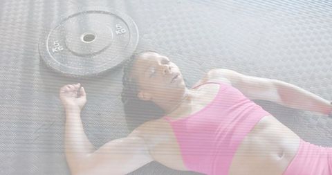 Woman lying on gym mat wearing pink sportswear after strength training with 5kg plate