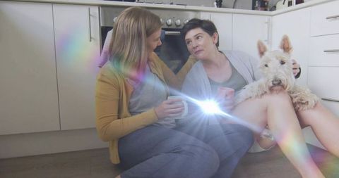 Two women sitting on kitchen floor sharing coffee with terrier and rainbow light flare