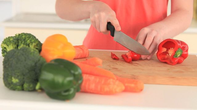 Hands hold knife slicing red bell pepper on wooden cutting board. Surrounding vegetables include vibrant broccoli, green pepper, yellow pepper, and carrots, suggesting focus on preparing fresh, healthy meals. Ideal used in promoting cooking blogs, health magazines, and nutrition-focused content.