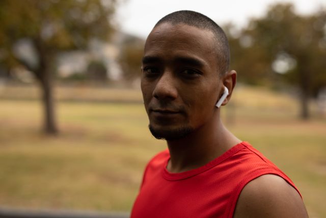 Biracial man wearing red sportswear and a smartwatch, listening to music on wireless earphones while taking a break from his running workout in a park. Ideal for use in fitness, health, and lifestyle promotions, as well as advertisements for sports gear, earphones, and wearable technology.