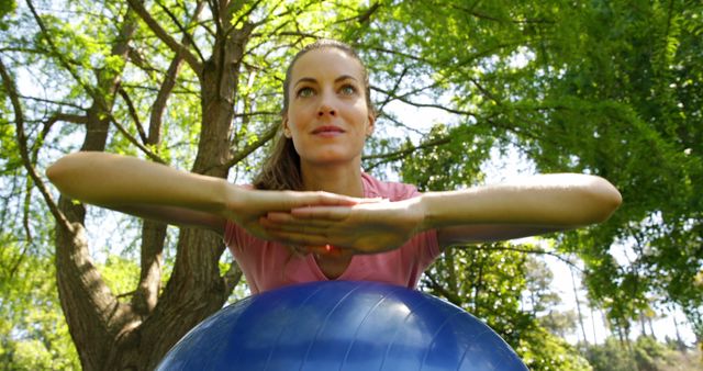 A woman is pressing arms down on a fitness ball outdoors in a lush, green park. She is engaging in a workout session focused on balance and wellness. This image can be used for promoting healthy living, fitness training programs, outdoor exercise routines, fitness equipment, and wellness blogs.