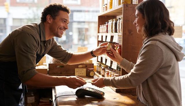 Smiling shopkeeper handing jar to customer at artisan market counter with card payment