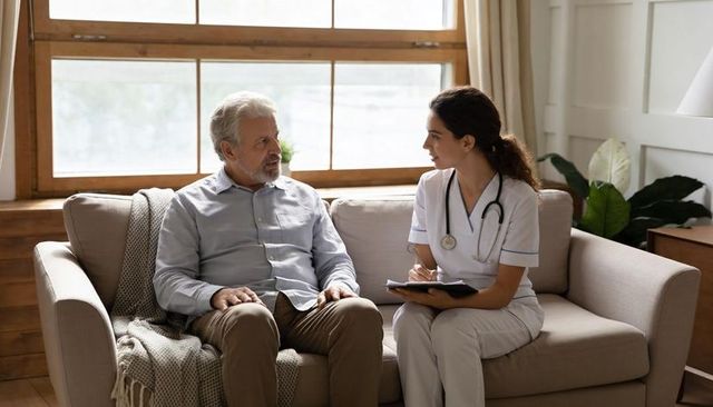 Nurse discussing care with senior man at home on sofa taking notes and assessing health
